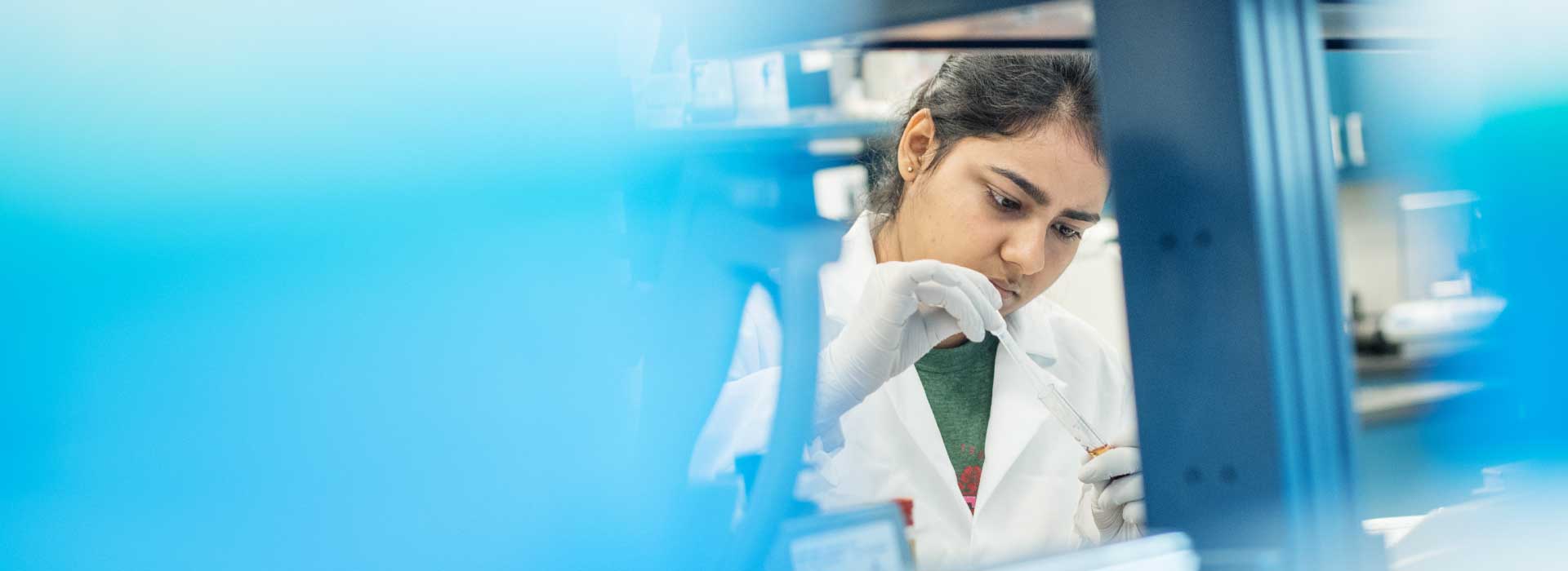 A woman in a research lab examines the contents of a test tube with gloved hands.