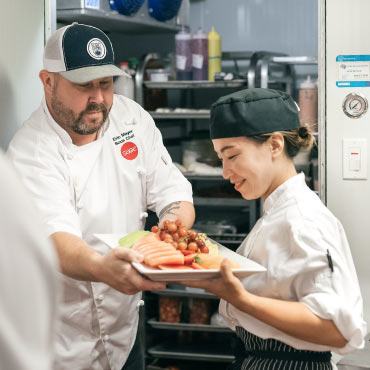 Two people working in an institutional kitchen holding a plate of fruit