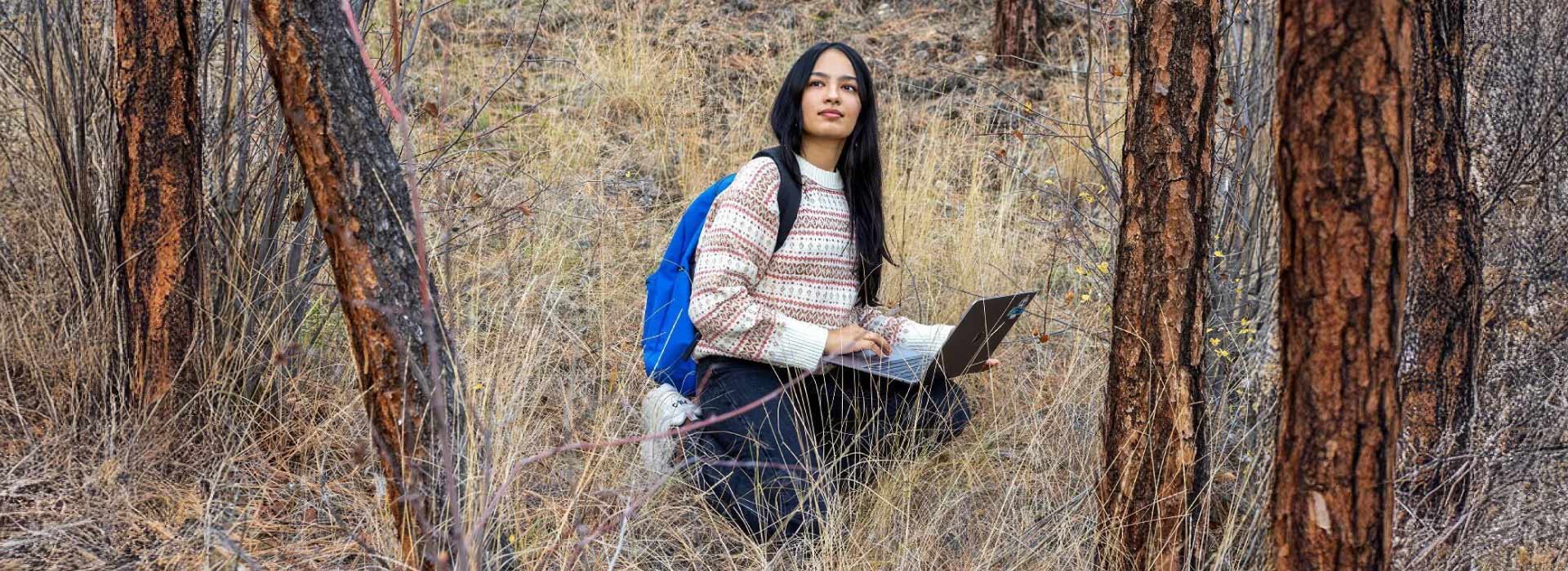 Shreya Saxena in a dry forest looking out into the distance, while kneeling down with her laptop on her lap