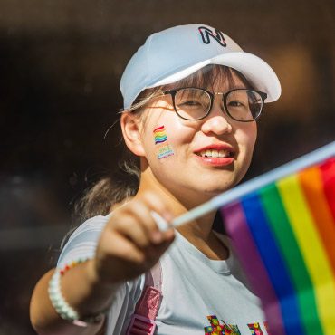 Person wearing glasses and a white cap smiles, holding a rainbow Pride flag. A rainbow sticker is on their cheek and sunlight illuminates their face.