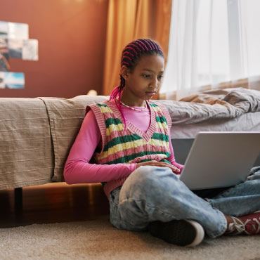 Youth on their laptop and sitting on the floor by bed
