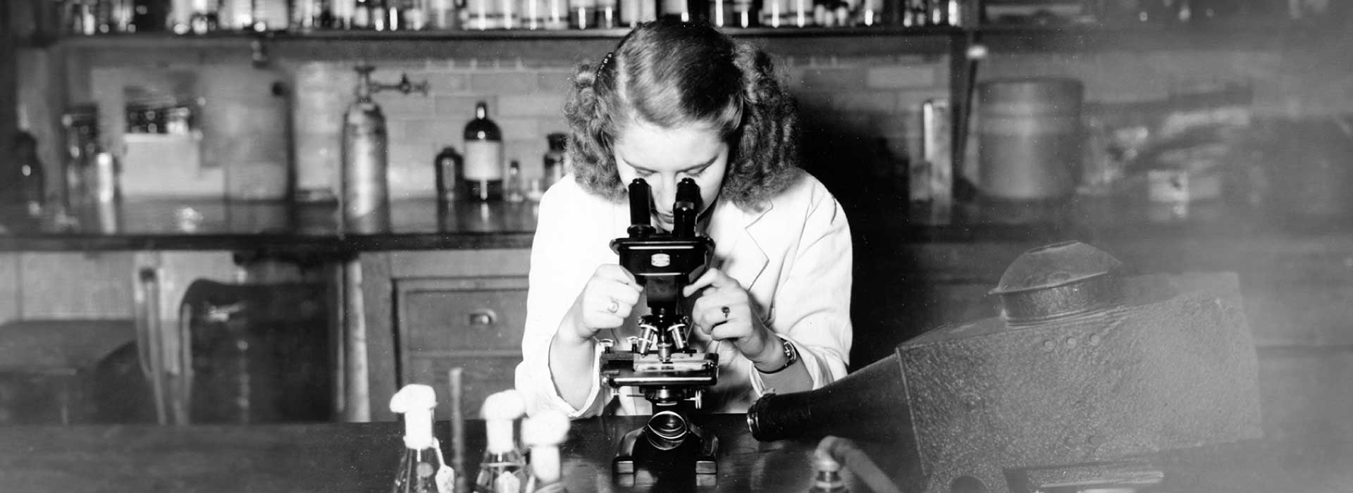 A woman in a lab coat looks through a microscope in a 1948 UBC Faculty of Medicine archival photo.